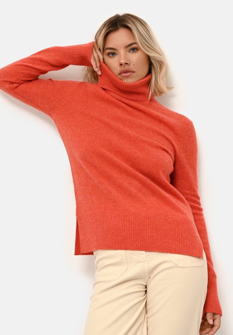 Maglione rosso a collo alto lavorato a maglia con dettagli a costine, caratterizzato da una vestibilità ampia e spacchi laterali. Abbinato a pantaloni beige chiaro.