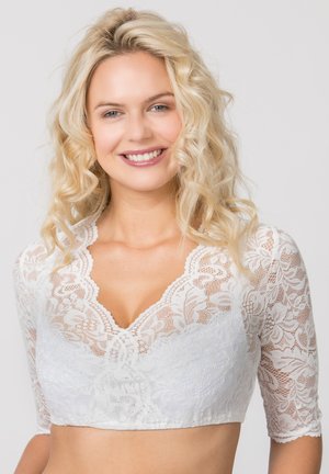 Blonde woman with curly hair smiling, wearing white lace cropped top with half sleeves against a plain light background.