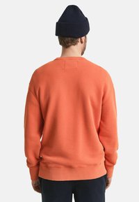 Oranje sweatshirt met geribbelde manchetten en zoom. Achterstevoren gedragen, heeft het een eenvoudig ontwerp en textielstof, gecombineerd met een donkere gebreide beanie.