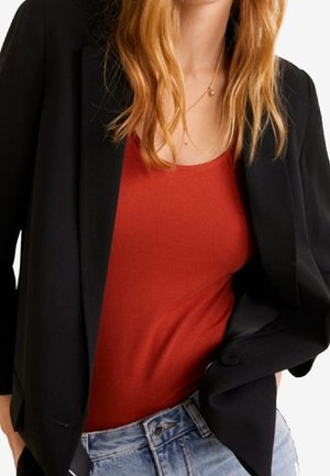 Femme portant un blazer noir par-dessus un haut rouge avec un jean bleu, montrant une tenue décontractée et stylée avec un délicat collier en or.