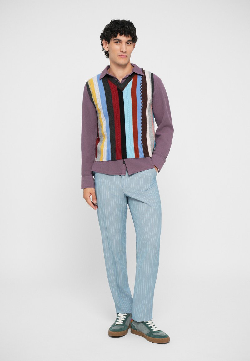 Gilet in maglia con strisce verticali multicolori, indossato sopra una maglietta a maniche lunghe viola. I pantaloni a righe sottili blu chiaro e le sneakers verdi completano l'outfit.