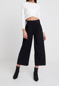Pantalon noir à jambes larges avec devant plissé et deux boutons à la ceinture, associé à un haut blanc court à manches longues.