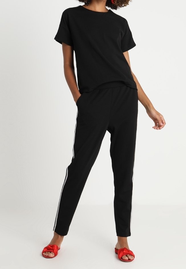 Personne portant une chemise noire à manches courtes et un pantalon noir avec des bandes blanches sur les côtés, debout les mains dans les poches, sandales rouges à bout ouvert.