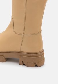 Béžová kožená boot s hladkým povrchom, masívnou hnedou gumovou podrážkou, spevneným šitím a mierne zaobleným tvarom špičky.