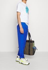 Bílý tričko s modrou grafikou, modré kalhoty a bílé tenisky s neonovými akcenty. Nese vícebarevnou tašku přes rameno s černými detaily.