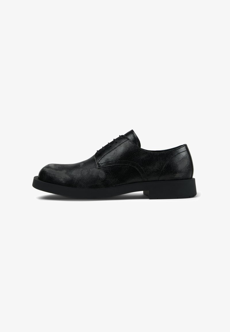 Zapato de vestir para hombre, negro de cuero con cordones, con una superficie ligeramente texturizada y una suela gruesa negra, mostrado de perfil sobre un fondo blanco.