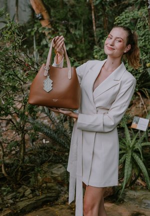 Braune Leder-Tote mit beigen Griffen, die ein dekoratives grünes Etikett und ein glänzendes Logo auf der Vorderseite aufweist. Glatte Textur, strukturierte Form.