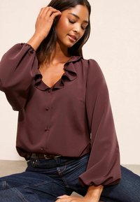 Mauvā blūze ar ruffles apkakli un aprocēm, ar pogu aizdari priekšā un brīvām piedurknēm, apvienota ar tumšajiem džinsiem.
