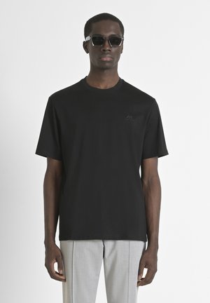 Maglietta nera a maniche corte con girocollo, con un logo discreto sul petto. Indossata con pantaloni grigi chiari e occhiali da sole.