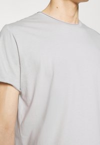 Buste masculin portant un t-shirt gris clair à manches courtes et col rond sur un fond uni.