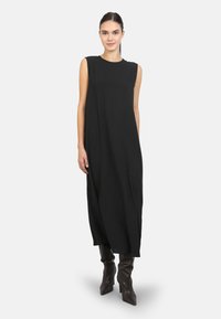 Schwarzes ärmelloses Kleid mit relaxed Fit, aus leichtem Stoff, mit Rundhalsausschnitt und bis zur Mitte der Waden reichend, kombiniert mit kniehohen Stiefeln.