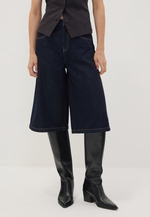 Persona que lleva culottes de mezclilla oscuros de pierna ancha, botas negras hasta la rodilla con tacón y un chaleco negro sin mangas y corto.