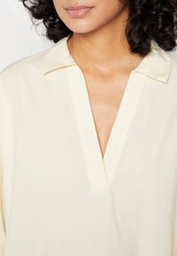Blouse jaune clair avec un col en V et un col pointu, confectionnée dans un tissu lisse, avec une coupe décontractée et des détails de couture minimaux.