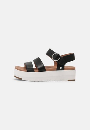 Sandale plateforme en cuir noir avec trois brides, une boucle ronde argentée et une semelle épaisse blanche avec une semelle marron.