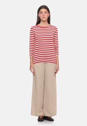 Donna in piedi di fronte, indossando una maglia a maniche lunghe a righe rosse e bianche, pantaloni beige a gamba larga e scarpe scure su sfondo bianco.