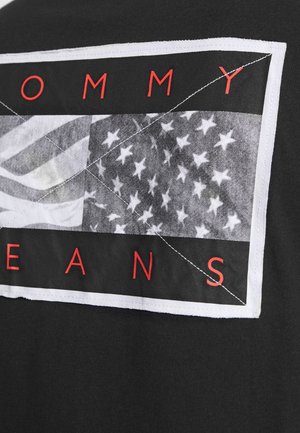 Black fabric with a stitched rectangular patch showing a black-and-white American flag and red letters spelling "TOMMY JEANS."