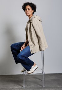 Beige lättviktsjacka med huva, denimbyxor med utsvängda ben och vita sneakers med beige detaljer, sittande på en genomskinlig stol mot en grå bakgrund.