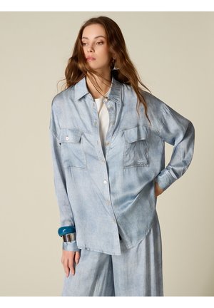 Frau mit langen braunen Haaren trägt ein lockeres helles Jeanshemd und passende Hose, ergänzt durch blaue und silberne Armbänder, schaut seitlich.