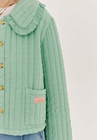 Světlá zelená prošívaná bunda s volánkovým límcem, dekorativním prošíváním a růžovou etiketou. Obsahuje velké knoflíky a přední kapsu.
