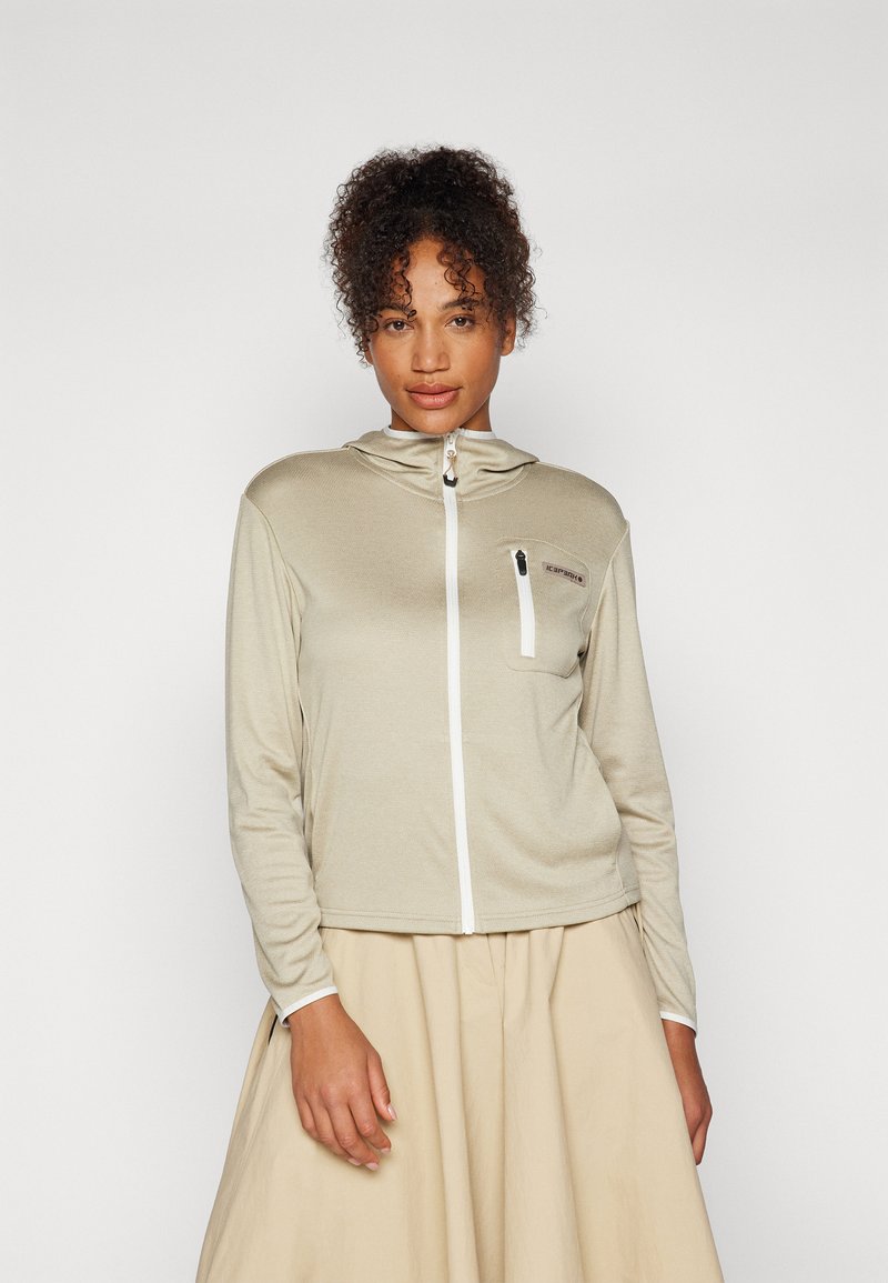 Lys beige zip-up hettegenser med lange ermer, teksturert stoff, frontlomme og hvite detaljer. Båret over et plisserte, lysfarget skjørt.