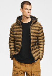 Brązowa kurtka puchowa o dopasowanym kroju, z poziomym pikowaniem, zamkiem błyskawicznym z przodu i kapturem. Noszona z czarną koszulą i beżowymi spodniami.