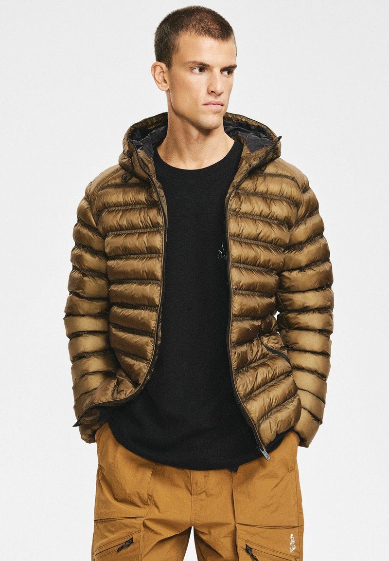 Brązowa kurtka puchowa o dopasowanym kroju, z poziomym pikowaniem, zamkiem błyskawicznym z przodu i kapturem. Noszona z czarną koszulą i beżowymi spodniami.