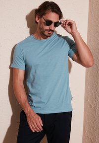 Tricou scurt, bleu deschis, dintr-un material moale și texturat. Are un guler rotund și o croială lejeră. Poartă pantaloni negri și ochelari de soare.