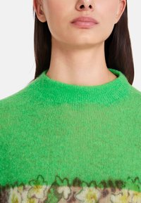 Grüner Strickpullover mit rundem Halsausschnitt, der am Saum ein Blumenmuster in gedämpften Farben aufweist. Weiche Textur, leicht durchscheinendes Aussehen.