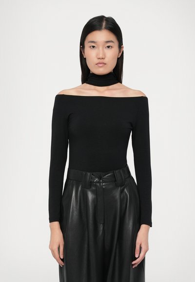 Giovane donna con capelli neri lisci indossa un top nero a spalle scoperte e pantaloni di pelle nera a vita alta su uno sfondo bianco semplice.