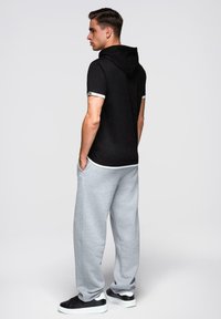 Mladý muž stojící bokem, oblečený v černé mikině s krátkým rukávem, světle šedých teplákách a černých teniskách s bílou podrážkou.