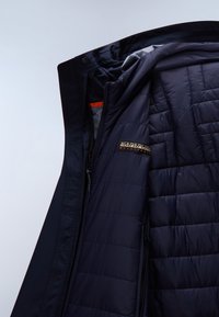 Marineblaue gesteppte Jacke geöffnet, um das Innenlogo-Patch und die Reißverschlusstasche vor einem schlichten hellen Hintergrund zu zeigen.