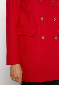 Manteau rouge croisé à boutons dorés, avec une poche latérale et un design légèrement structuré. Le tissu semble lisse et chaud.