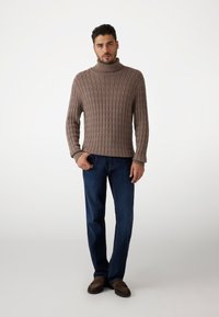 Bruine gebreide trui met hoge hals, gecombineerd met donkerblauwe rechttoe rechtaan jeans en bruine instap schoenen, tegen een lichte achtergrond.
