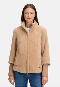 Kobieta ubrana w beżową, futrzaną kurtkę z zamkiem błyskawicznym, pod którą ma na sobie prążkowaną bluzkę z długim rękawem oraz niebieskie dżinsy, stoi na jednolitym tle.