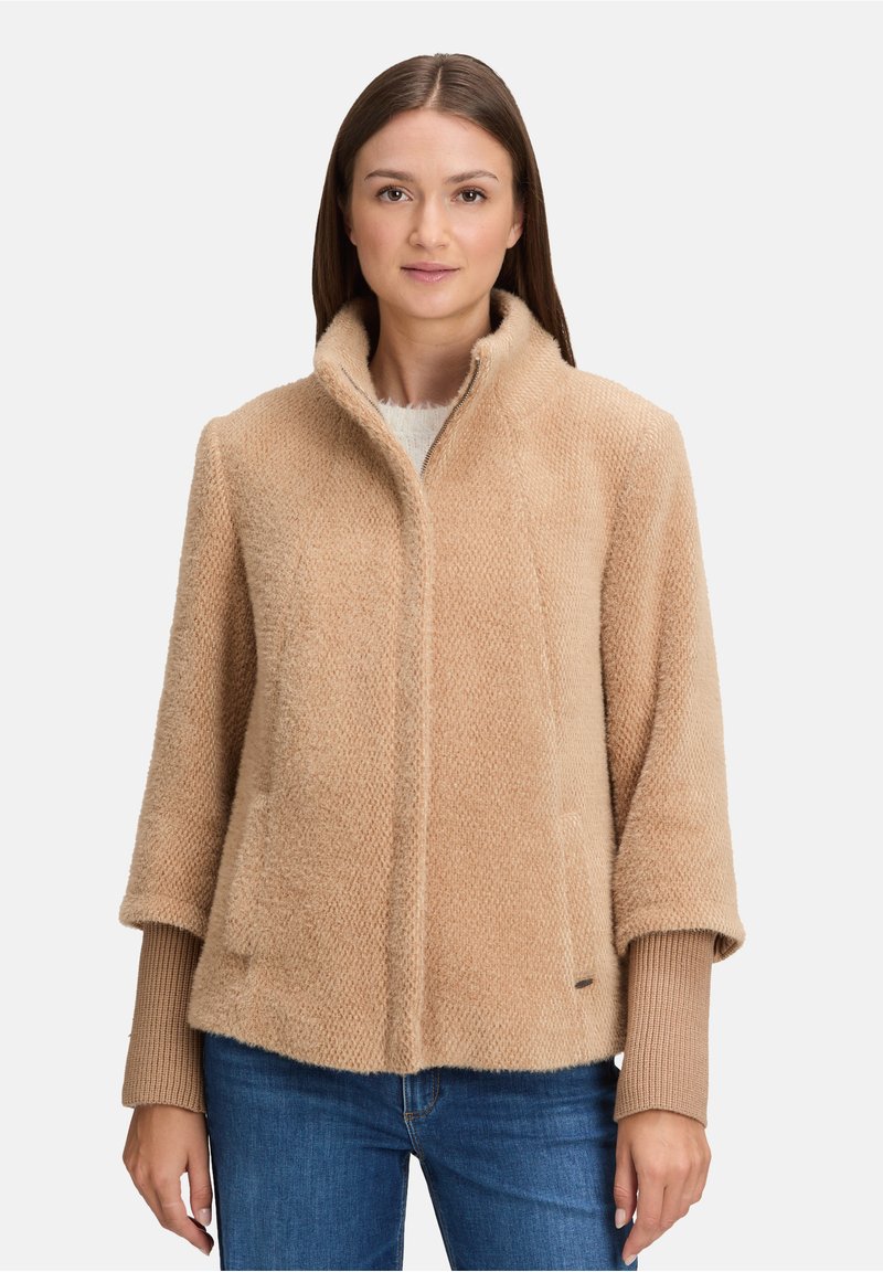 Kobieta ubrana w beżową, futrzaną kurtkę z zamkiem błyskawicznym, pod którą ma na sobie prążkowaną bluzkę z długim rękawem oraz niebieskie dżinsy, stoi na jednolitym tle.