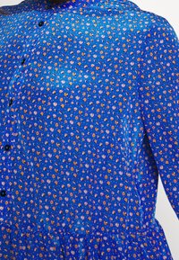 Blå mönstrad blus i ett mjukt tyg. Har små blommönster i gult och vitt, med svarta knappar fram och en rundad krage.