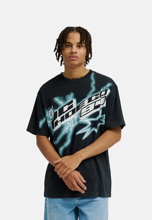 Jeune personne avec de courtes tresses se tenant face à l’avant, portant un t-shirt noir à motif graphique avec des éclairs turquoise et un texte blanc en gras, et un jean bleu clair.