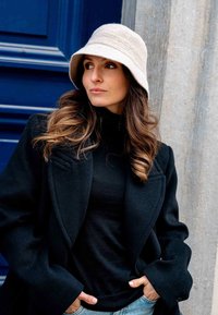 Felt bucket hat in light beige, black wool coat with tailored lapels, fitted black top, and blue jeans, against a blue door backdrop.