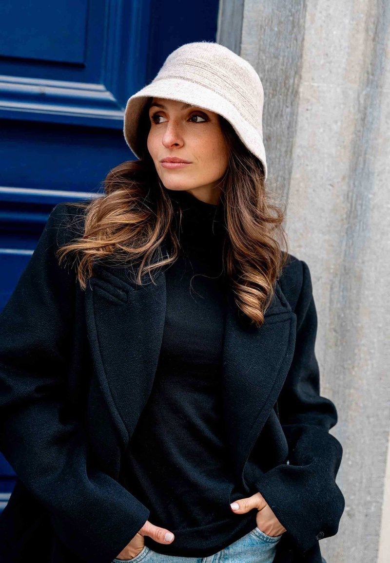 Felt bucket hat in light beige, black wool coat with tailored lapels, fitted black top, and blue jeans, against a blue door backdrop.