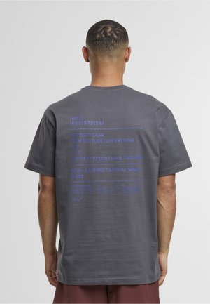 Uomo di spalle che indossa una maglietta grigia con testo stampato blu sul retro che fa riferimento a termini militari e di allenamento fisico.