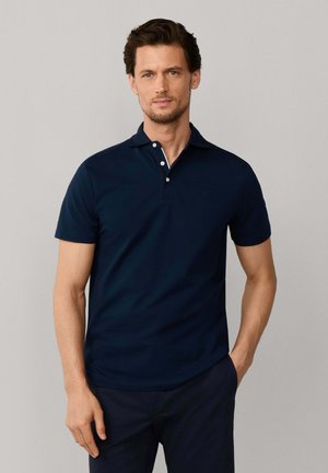 Man met kort bruin haar, gekleed in een donkerblauw poloshirt met korte mouwen en bijpassende broek, staand met één hand in de zak.