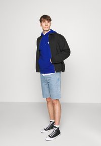 Giacca nera sopra un felpa blu, con pantaloni shorts in denim blu chiaro e scarpe alte nere. Design semplice senza motivi visibili.