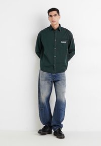 Dark green button-up shirt with a chest pocket, paired with light blue distressed denim jeans and black shoes, set against a white background.