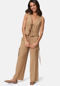 Tan, ärmlös jumpsuit med djup V-ringning, snörning i midjan och cargo-fickor. Mjuk tyg med slät textur, bärs med nitar på ballerina.