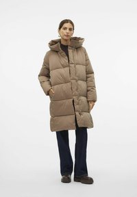 Abrigo marrón acolchado con capucha, que cuenta con un cierre de cremallera, bolsillos laterales y un corte hasta la rodilla, llevado sobre pantalones oscuros y zapatos.