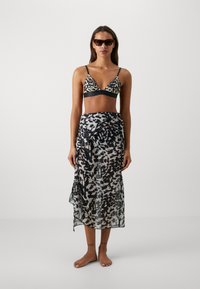 Zwart en beige luipaardprint bikini-top met dunne straps, gedragen met een doorschijnende zwarte en grijs gemusterde wikkelrok. Model staat op blote voeten.