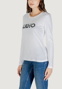 Modello in camicia bianca a maniche lunghe con "LIUJO" in nero sul petto, abbinata a jeans blu, posato su uno sfondo neutro.