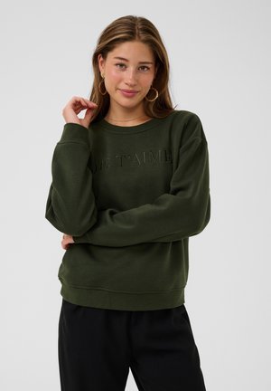 Jeune femme aux cheveux châtain clair portant des boucles d’oreilles en anneaux dorés et un sweat-shirt vert foncé avec le texte "JE T'AIME", posant avec la main près du visage.
