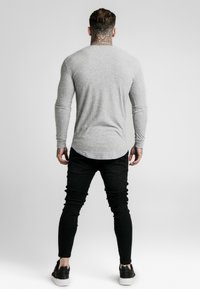 Mann steht mit dem Rücken zur Kamera, trägt ein hellgraues geripptes Langarmshirt, schwarze enge Jeans und schwarze Sneaker mit weißen Sohlen.