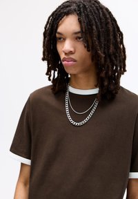 Brązowy T-shirt z białym wykończeniem rękawów, o luźnym kroju. Dwie srebrne łańcuszki opadają na szyję, dopełniając całość.